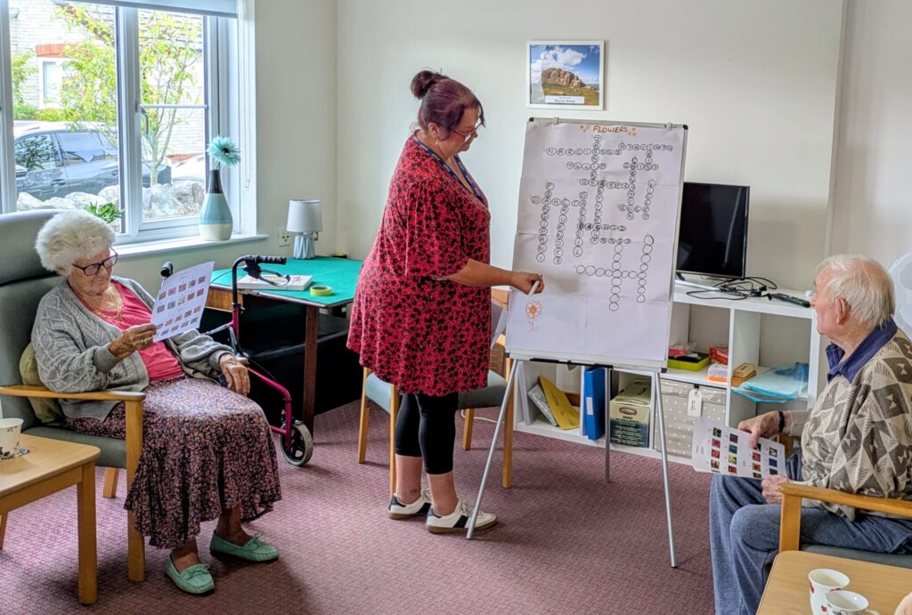 Care home activity: a staff member presents a word puzzle on a flip chart as three elderly residents watch.