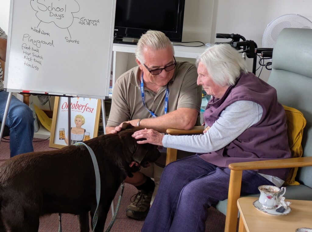 Two elderly people sit together, one stroking a large dark dog in a cozy room with a whiteboard nearby showing a 'School Days' diagram.