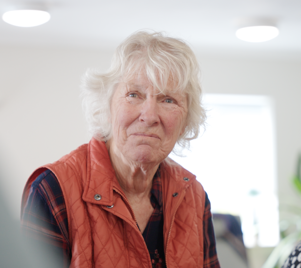 Close-up of an elderly woman with short white hair wearing an orange quilted vest, indoors.