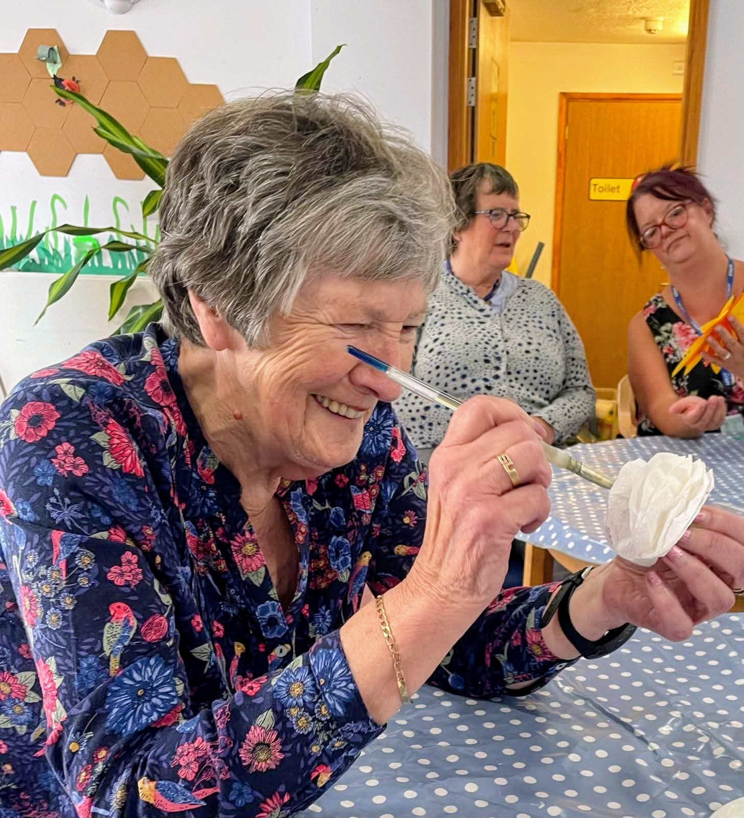 Smiling elderly woman with short gray hair in a blue floral blouse holds a blue pen near her nose at a table with a polka-dot tablecloth.