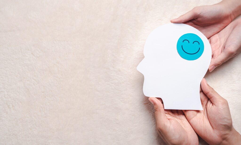 Hands hold a white paper head silhouette with a blue smiling circle sticker on the cheek, against a beige textured background.